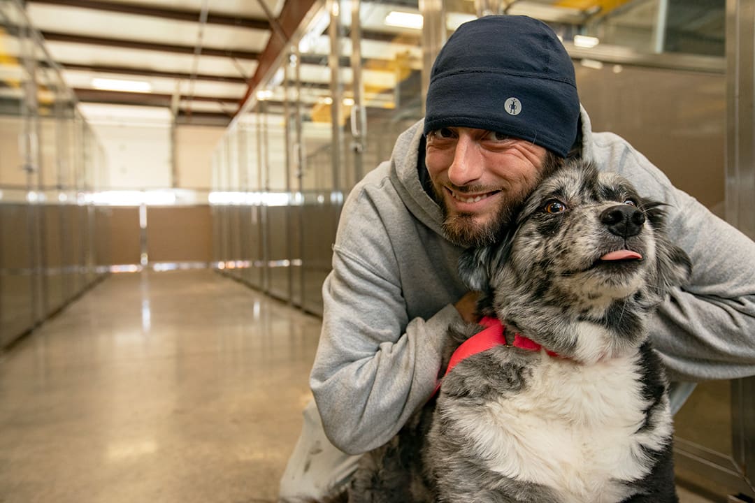 A male Vera's Posh Employee smiles while hugging a happy dog at daycare.